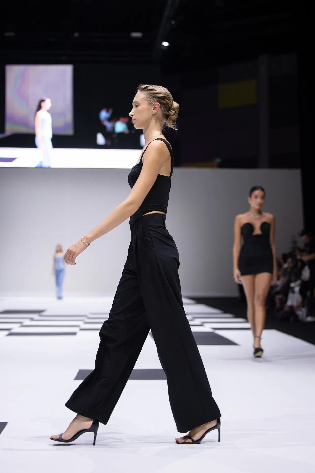 Model walking the runway in a black sleeveless top and wide-leg pants, wearing strappy heels at a fashion show.