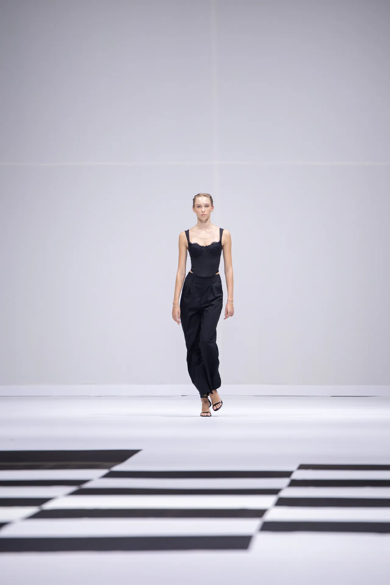 Female model in a black sleeveless top and black trousers walking down a minimalist runway with a geometric black-and-white floor pattern.