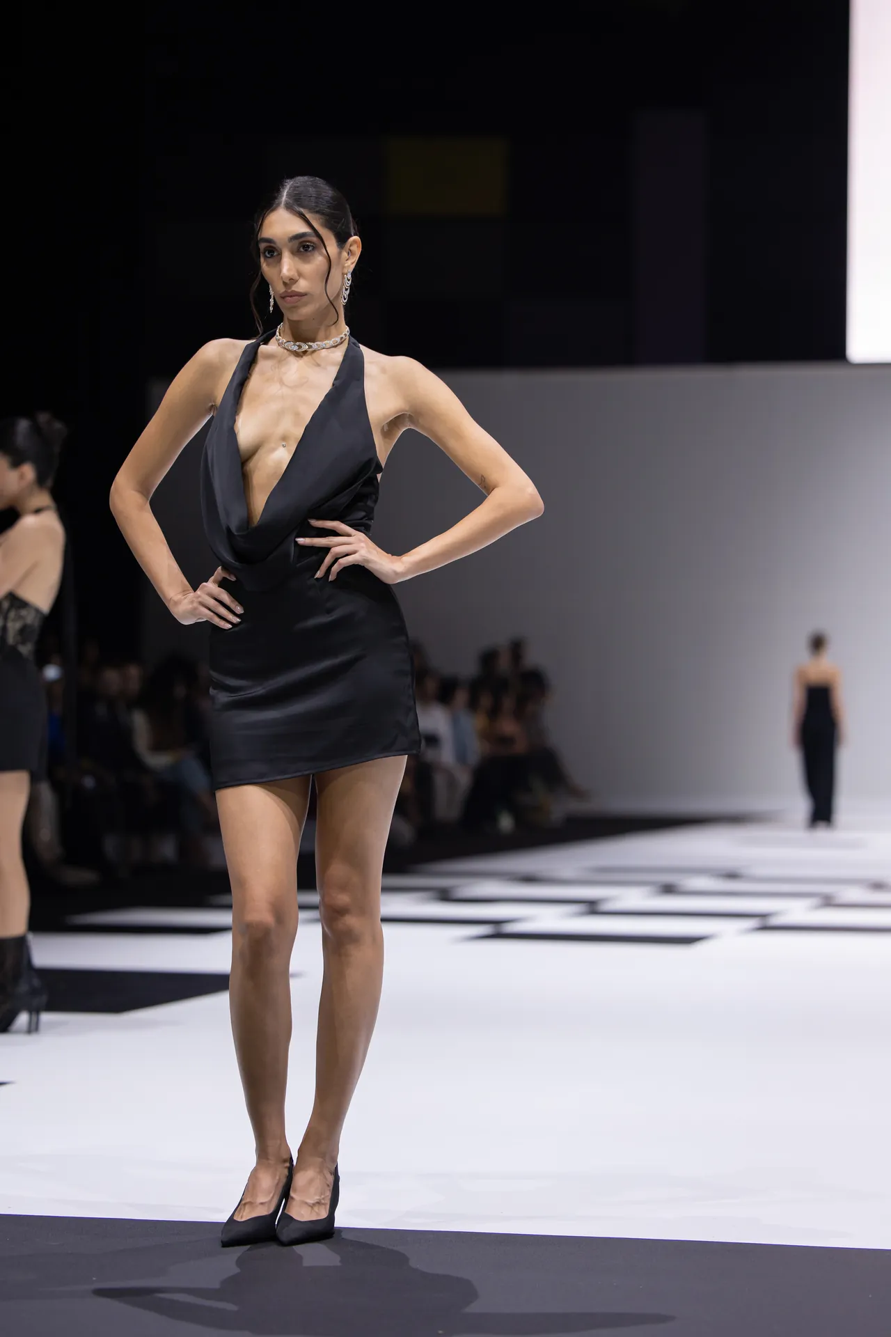 Model on a runway wearing a black halter-neck mini dress and black heels, posing with hands on hips.