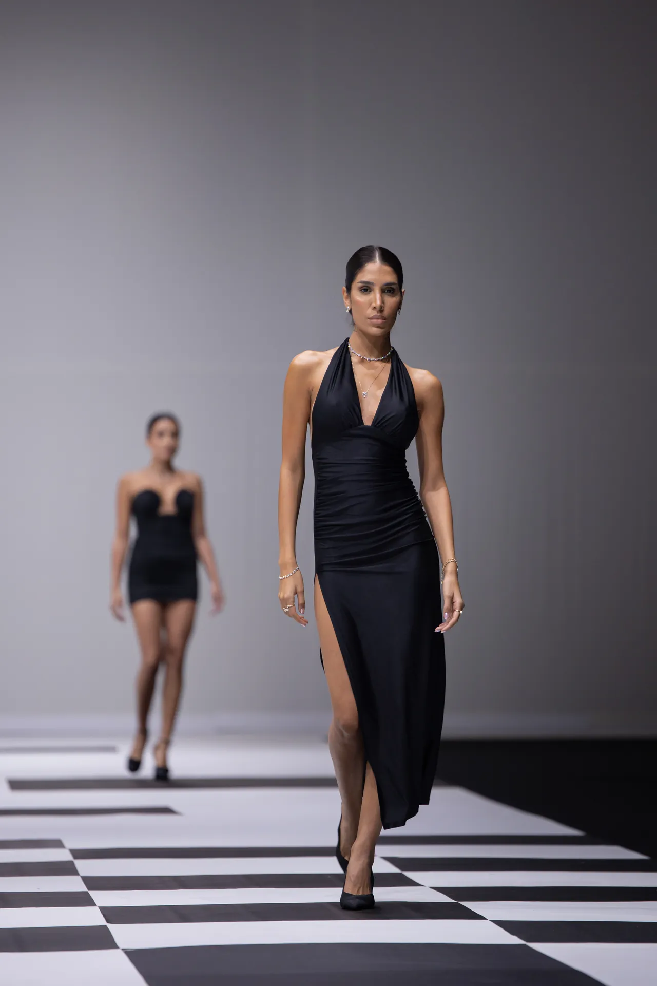 Model in a black halter gown walking toward the camera on a bold black-and-white checkerboard runway; background model blurred.