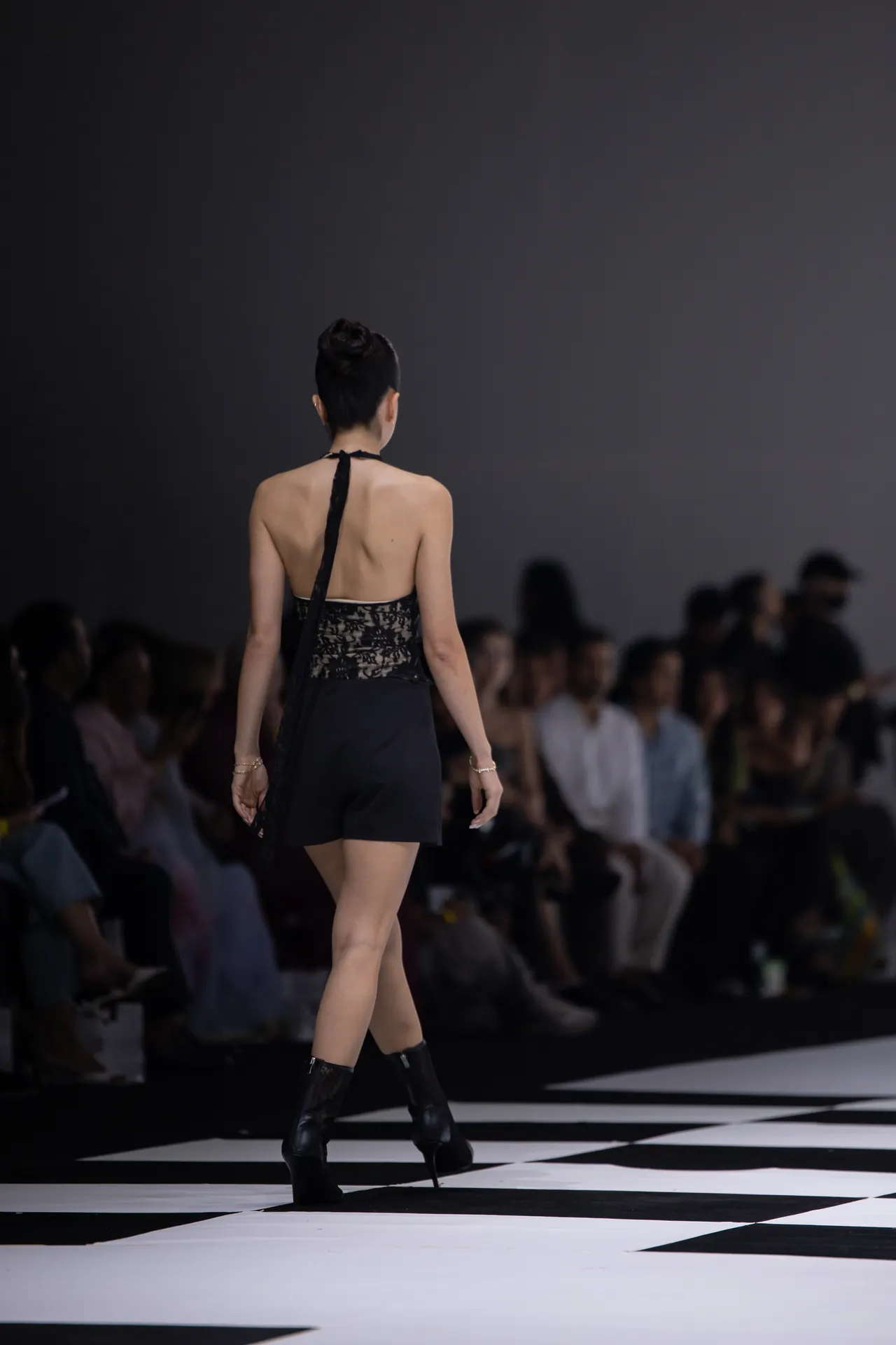 Back view of a fashion model walking on a black-and-white checkered runway, wearing a black lace halter top and shorts.