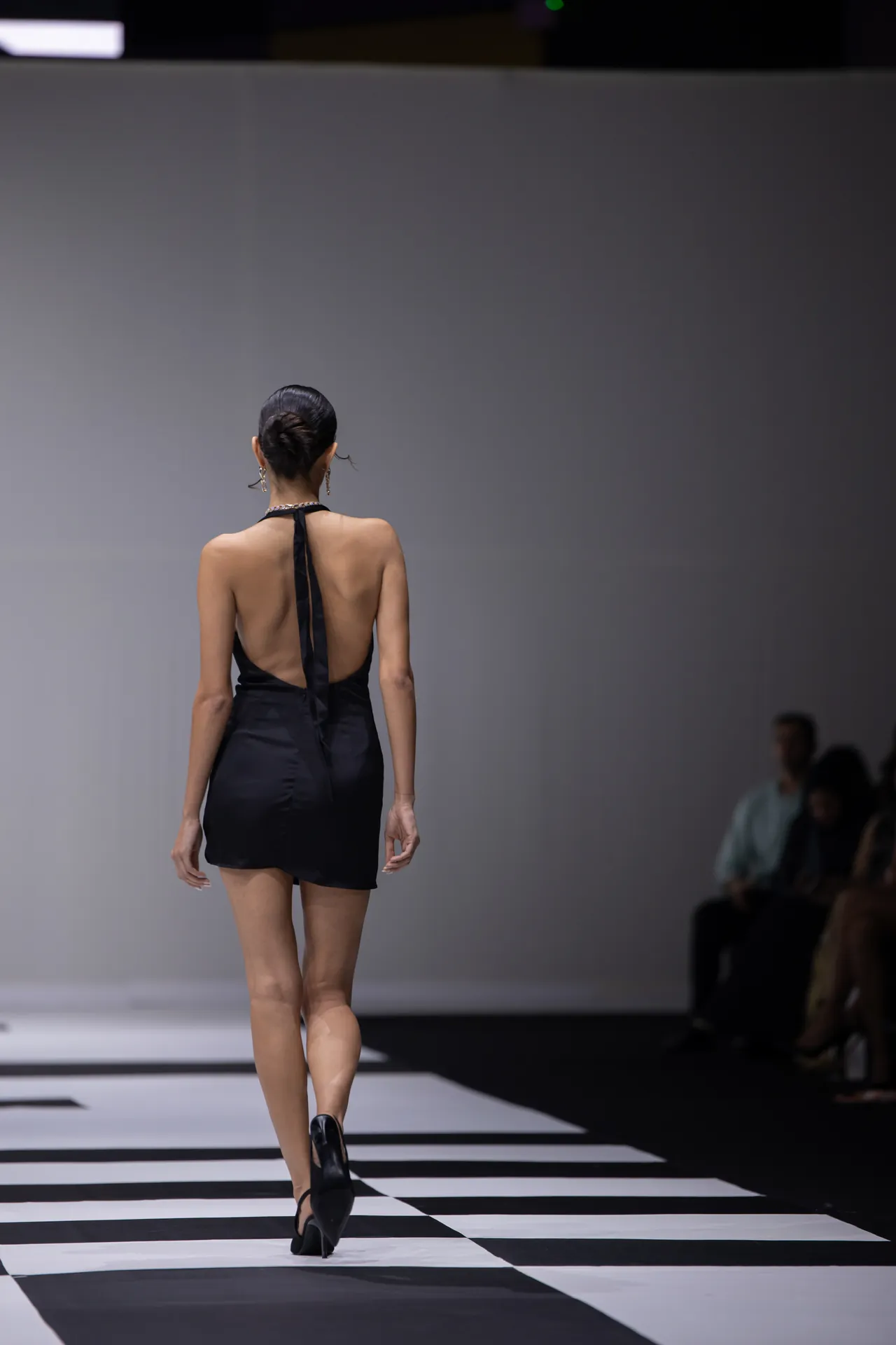 Model wearing a backless black dress walking away on a black-and-white striped runway.