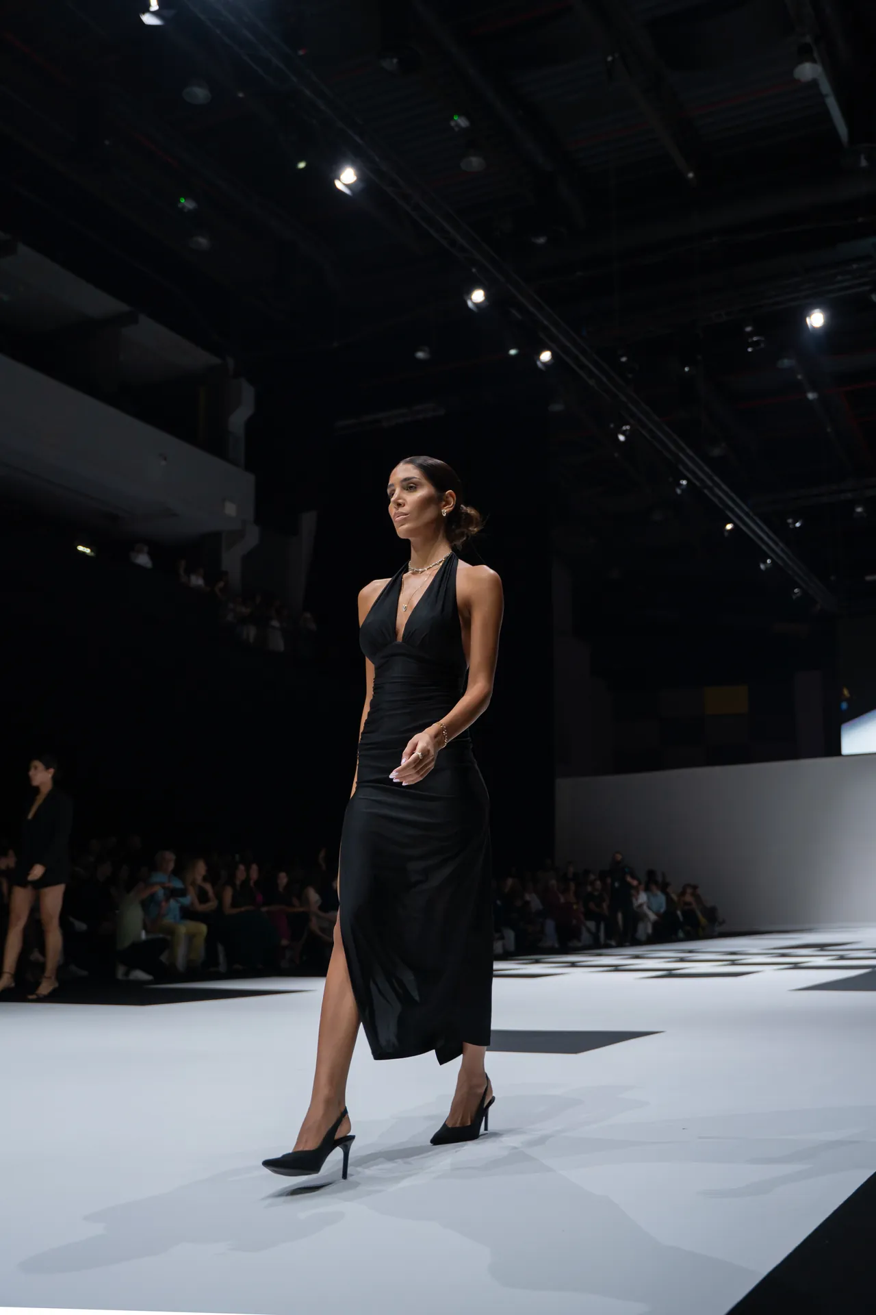 Model in a black halter gown walking down a fashion show runway under bright stage lights.