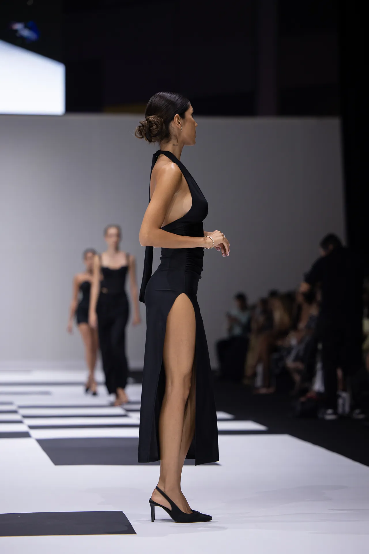 Profile view of a female model walking a black halter gown with a high slit on a fashion runway, audience visible in the background.