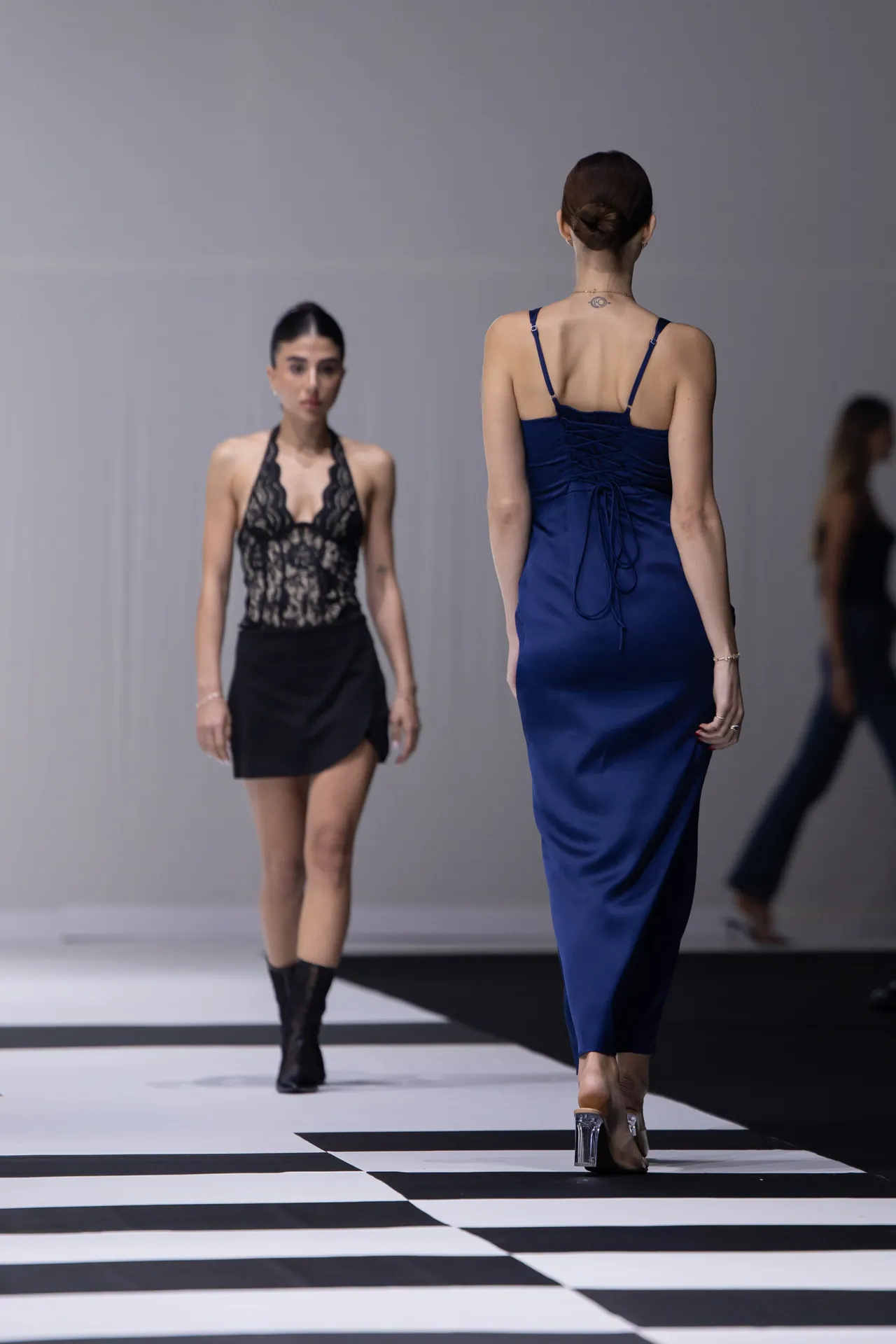 Model walking a fashion show in a blue satin dress seen from the back, with a second model in a black lace dress approaching on a black-and-white runway.
