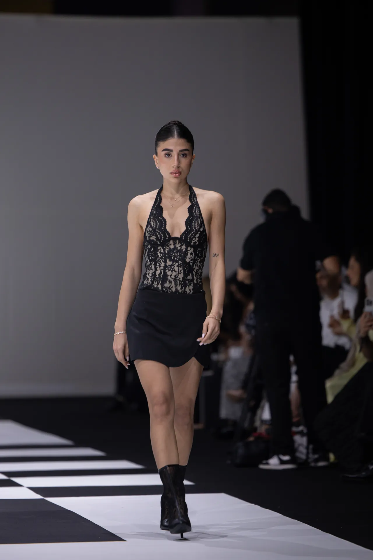 Model walks down a fashion runway wearing a black lace halter top and black mini skirt.