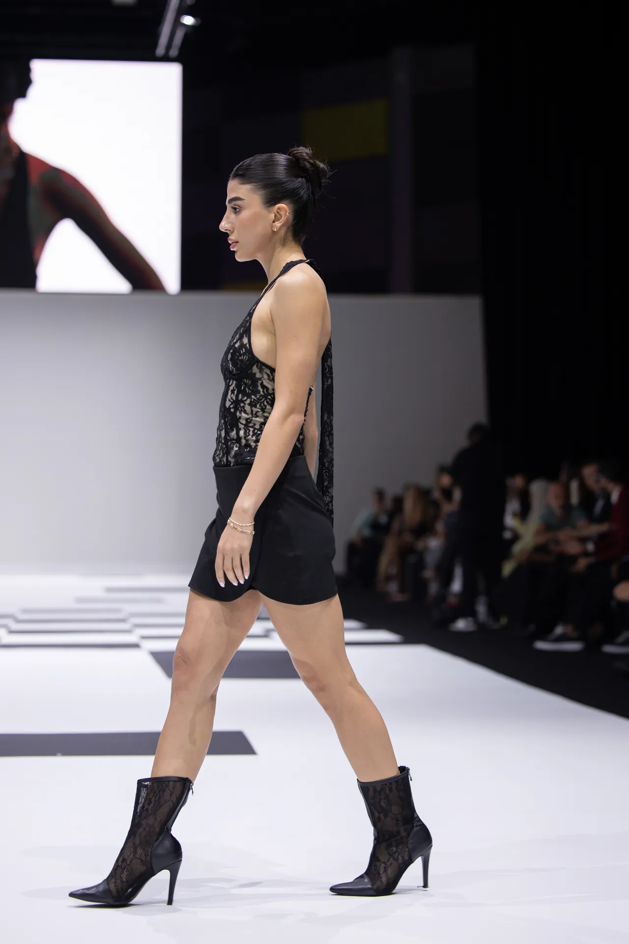 Model walking on a runway in a black lace halter top, black shorts, and lace boots, side profile view.