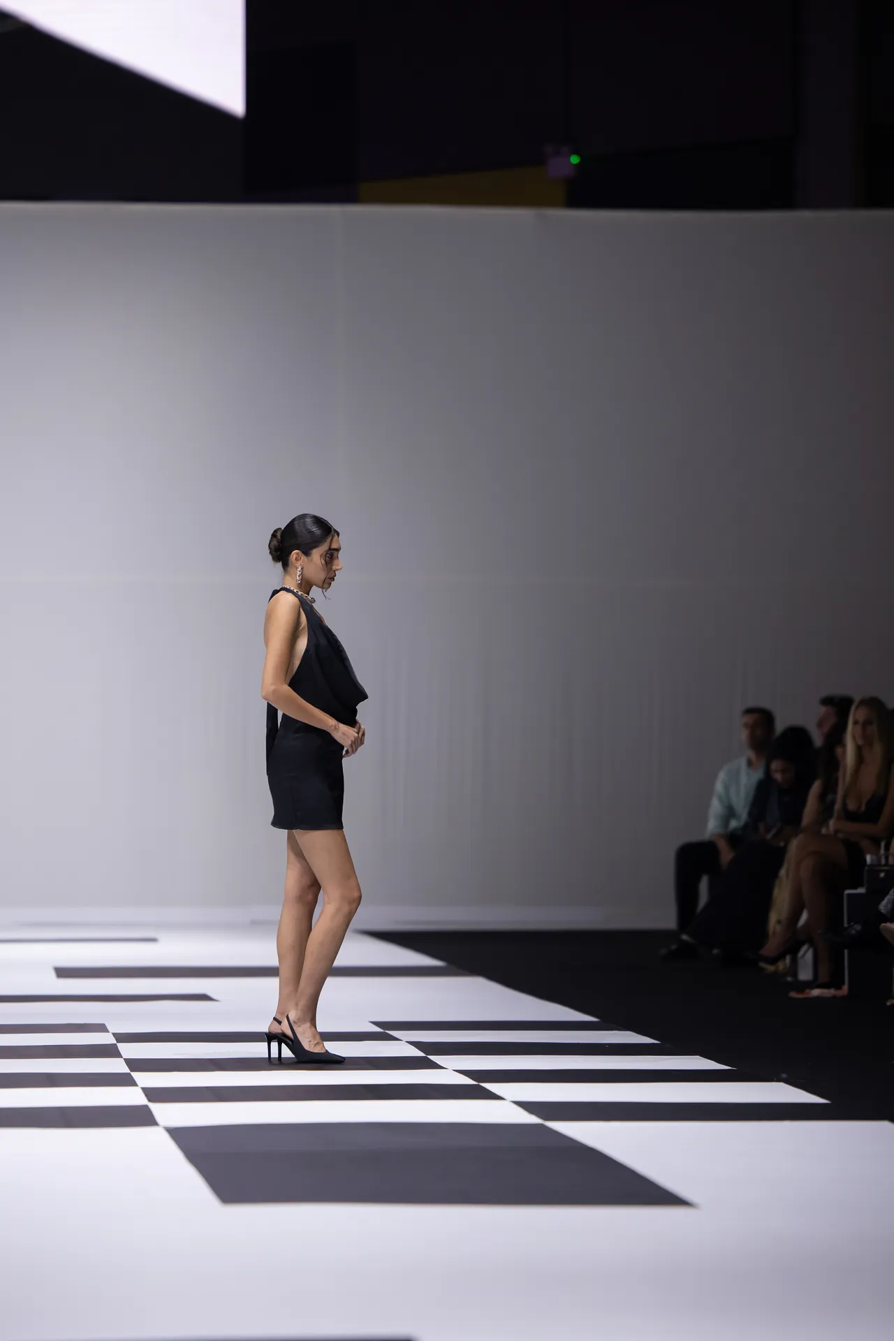 Female model in a black sleeveless dress walking a black-and-white checkered runway; audience seated along the wall.