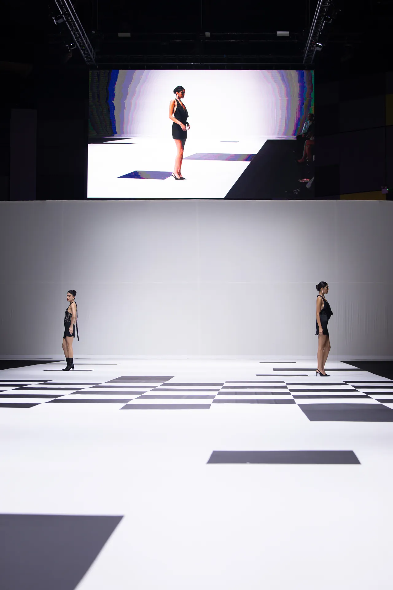 Runway show: three models stand apart on a white stage with black geometric floor patterns; a large screen behind them displays another model in a black dress.