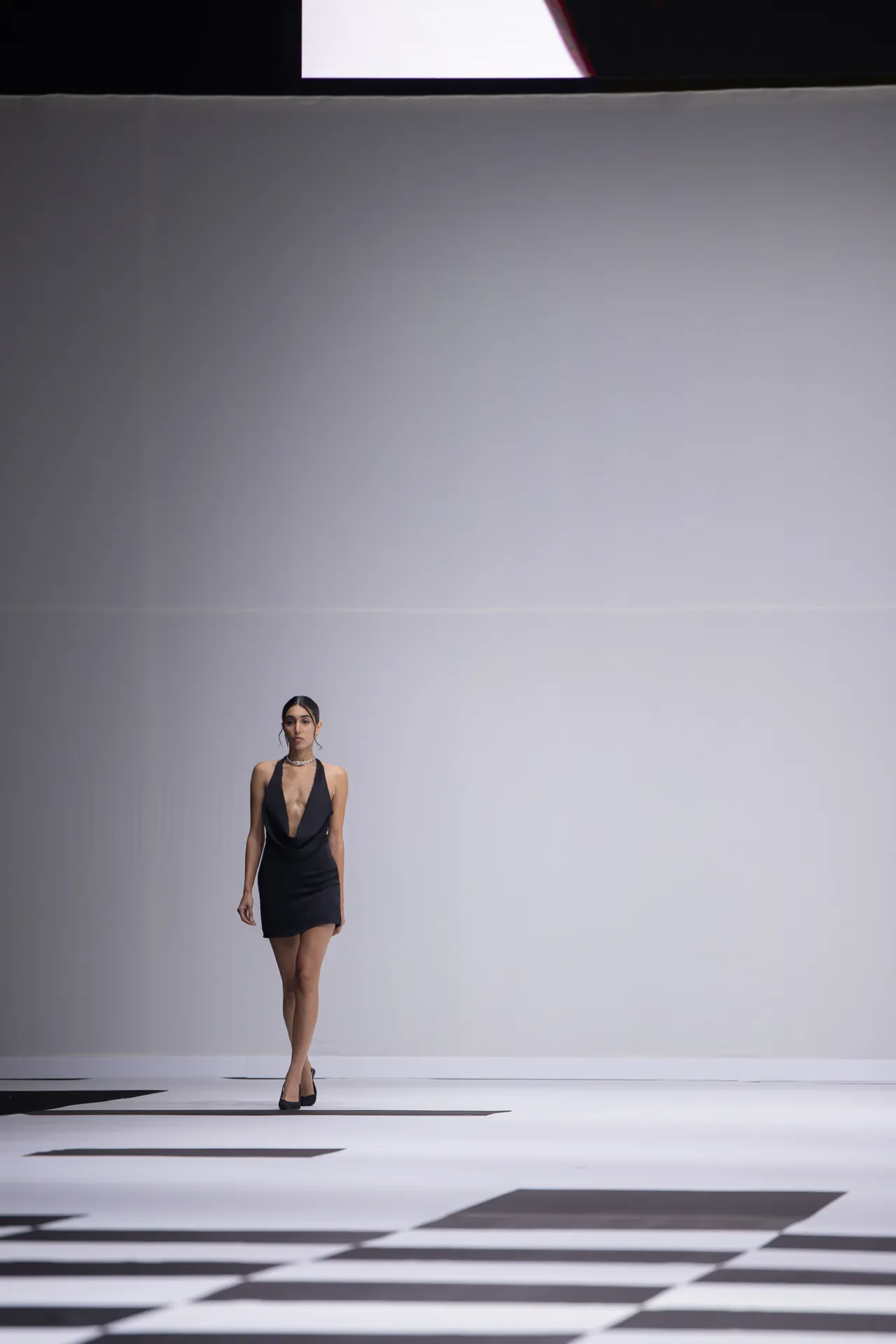 Model walking a runway in a black halter dress against a minimalist gray backdrop; geometric black-and-white floor in foreground