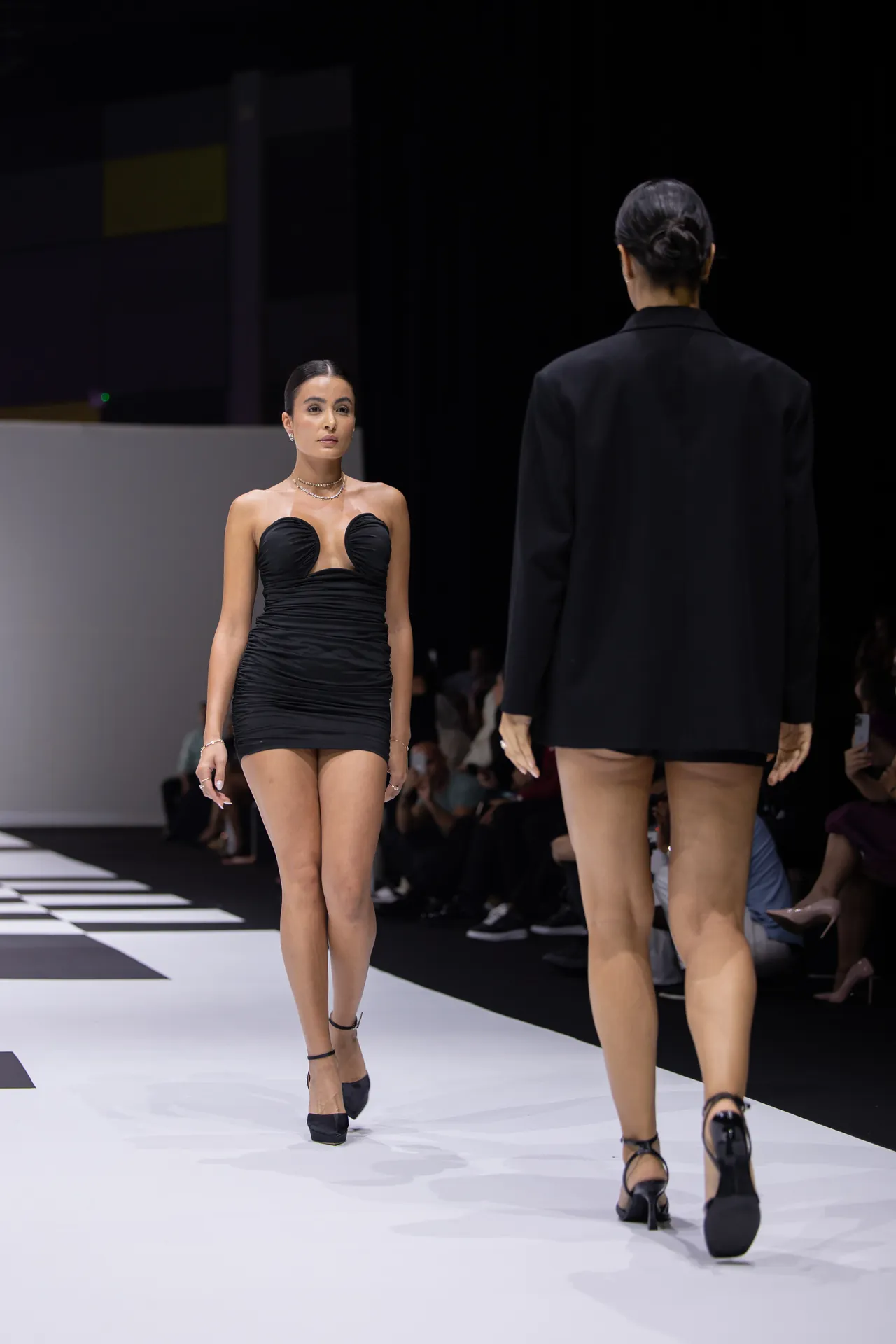 Runway scene with a woman in a black strapless mini dress walking toward the camera as another model in a black blazer walks away down the catwalk; audience visible in the background.