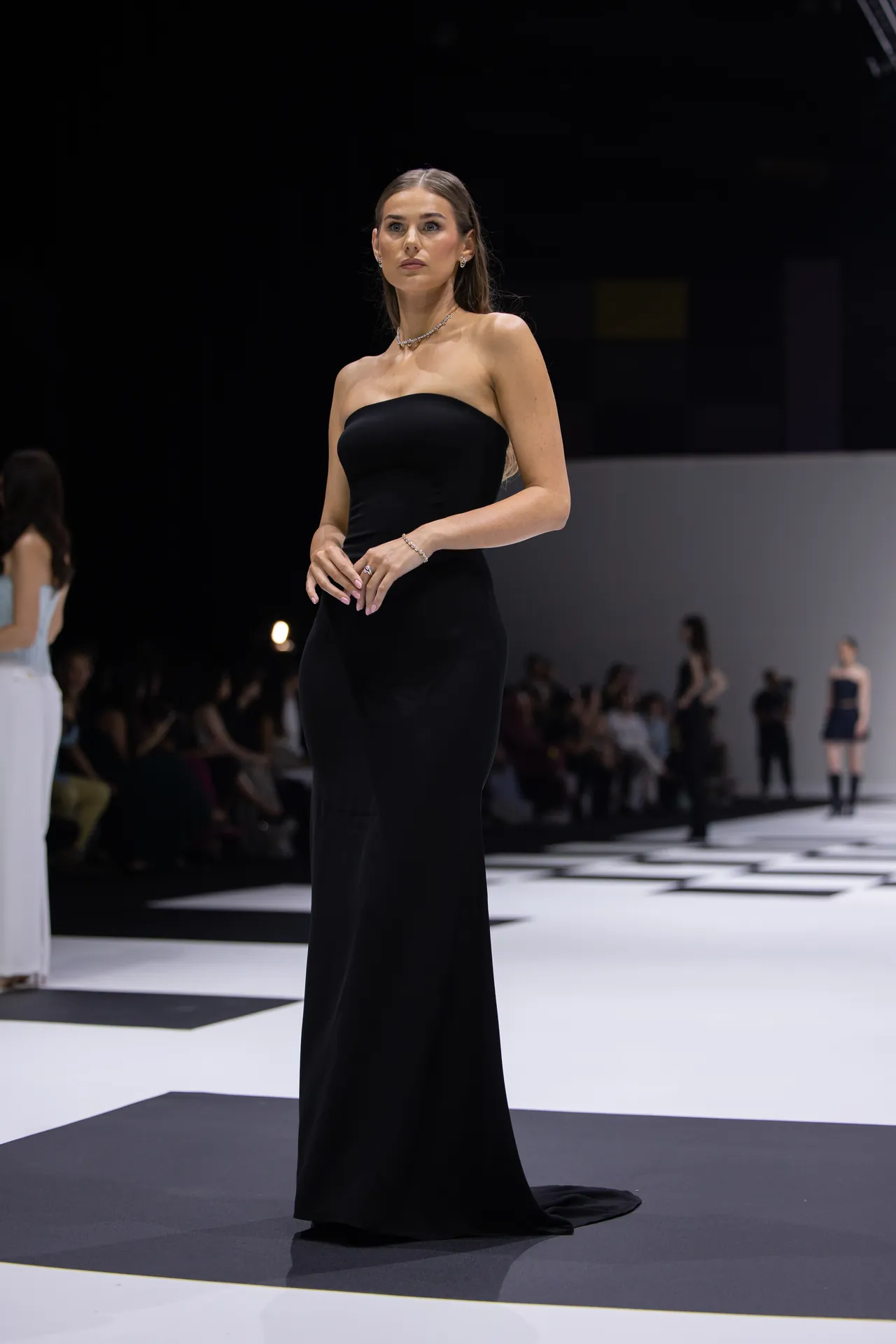 Model in a strapless black evening gown posing on a runway; audience blurred in the background.