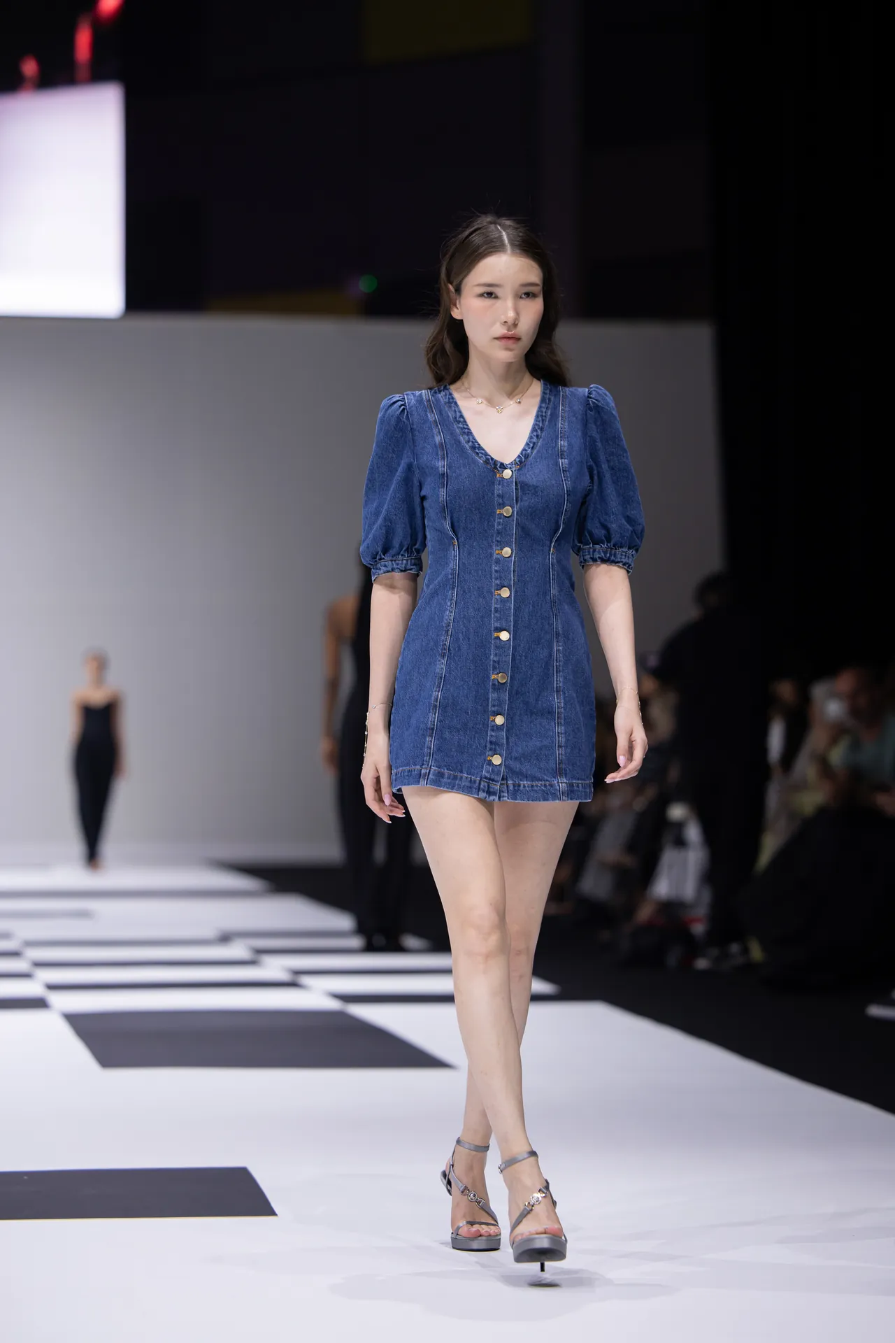 Model walking a fashion runway in a blue denim button-front mini dress with puff sleeves and sandals.
