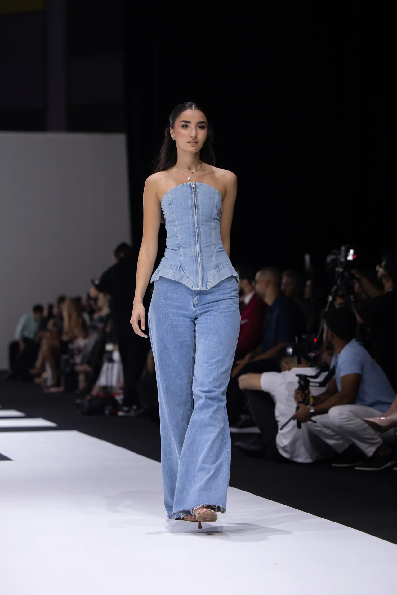 Model walks the runway in a strapless light denim peplum top and wide-leg jeans, photographers in the background.
