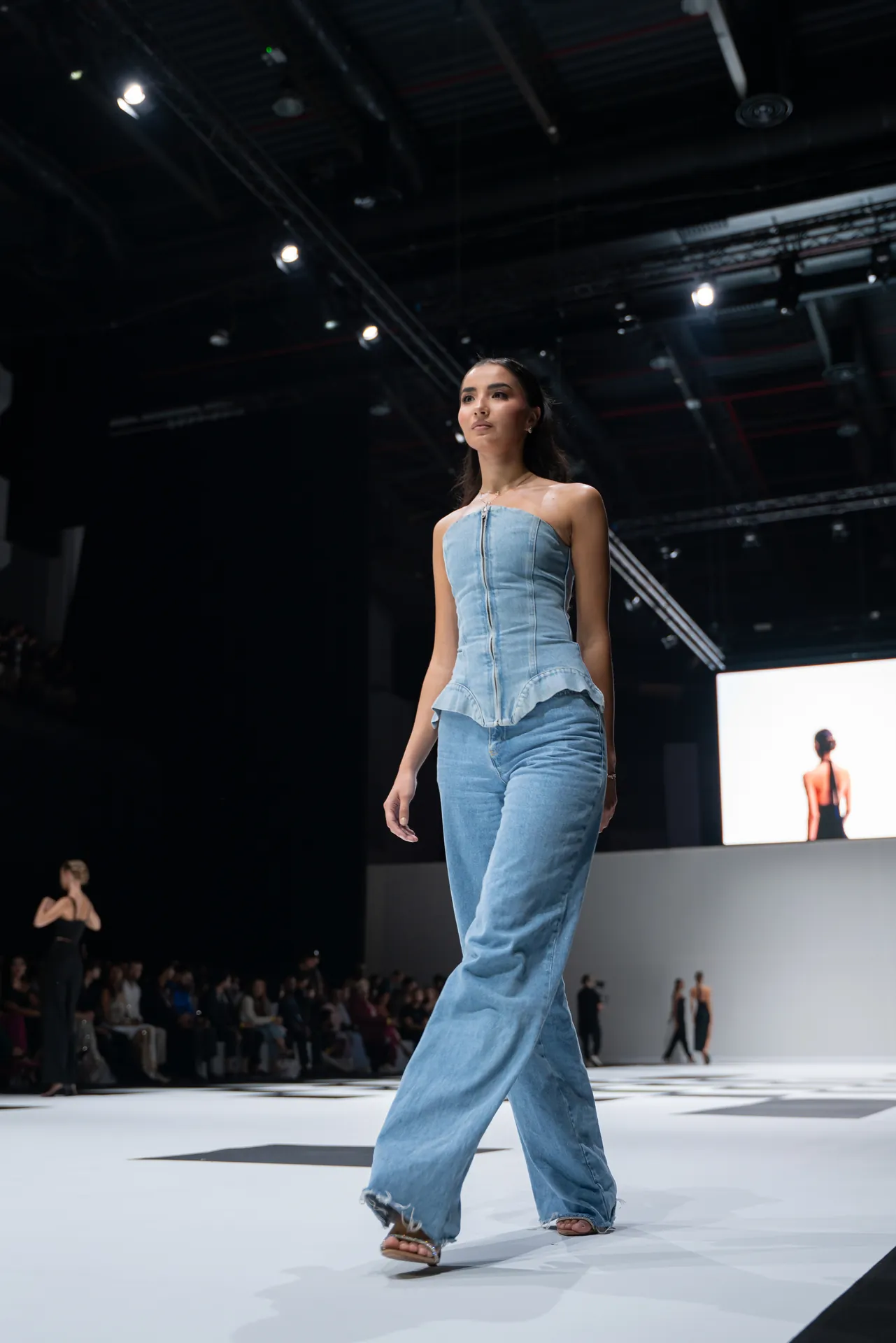 Female model walks the runway in a light denim strapless top and wide-leg jeans on a fashion show catwalk.