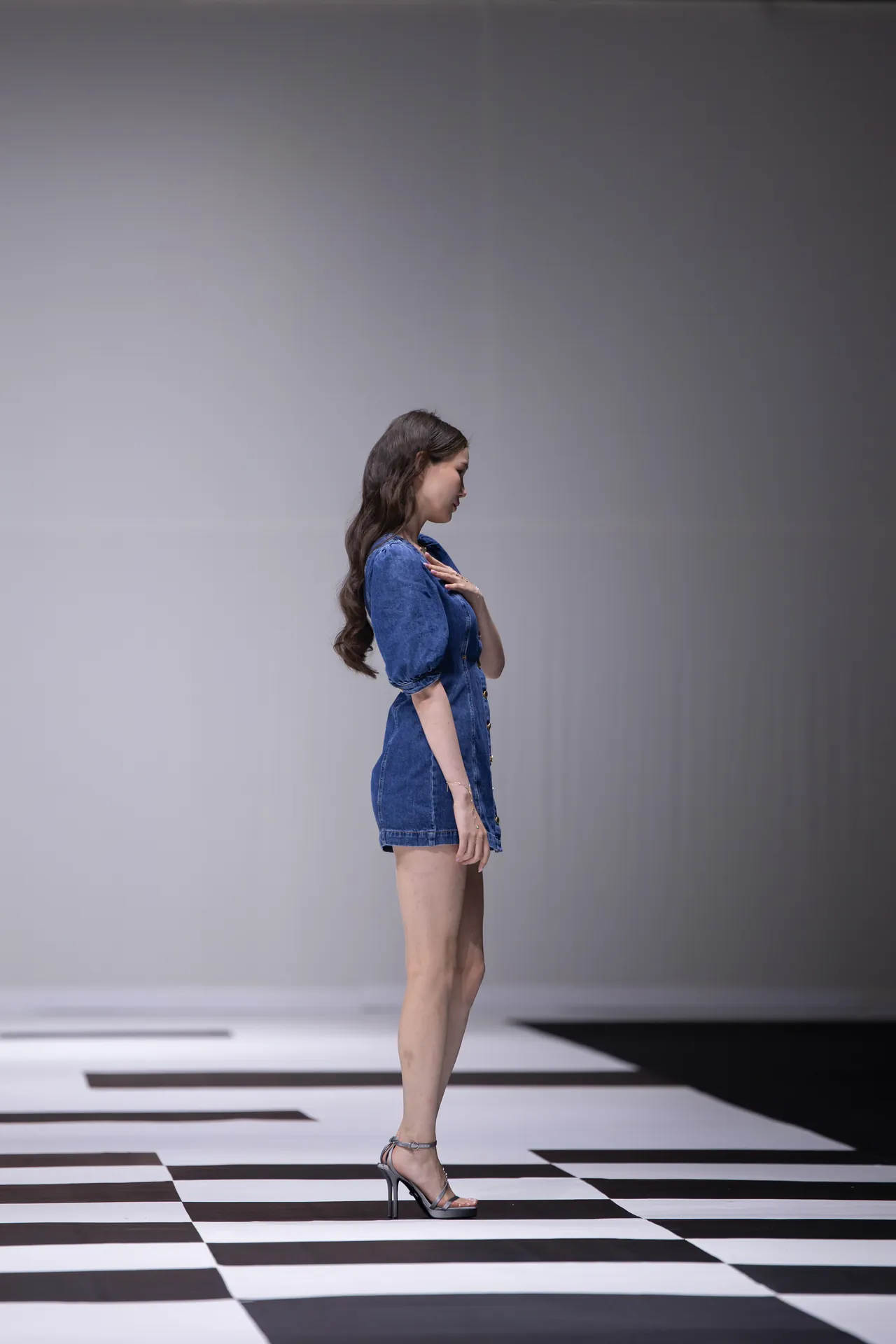 Side view of a woman in a blue denim dress walking on a black-and-white checkerboard runway.
