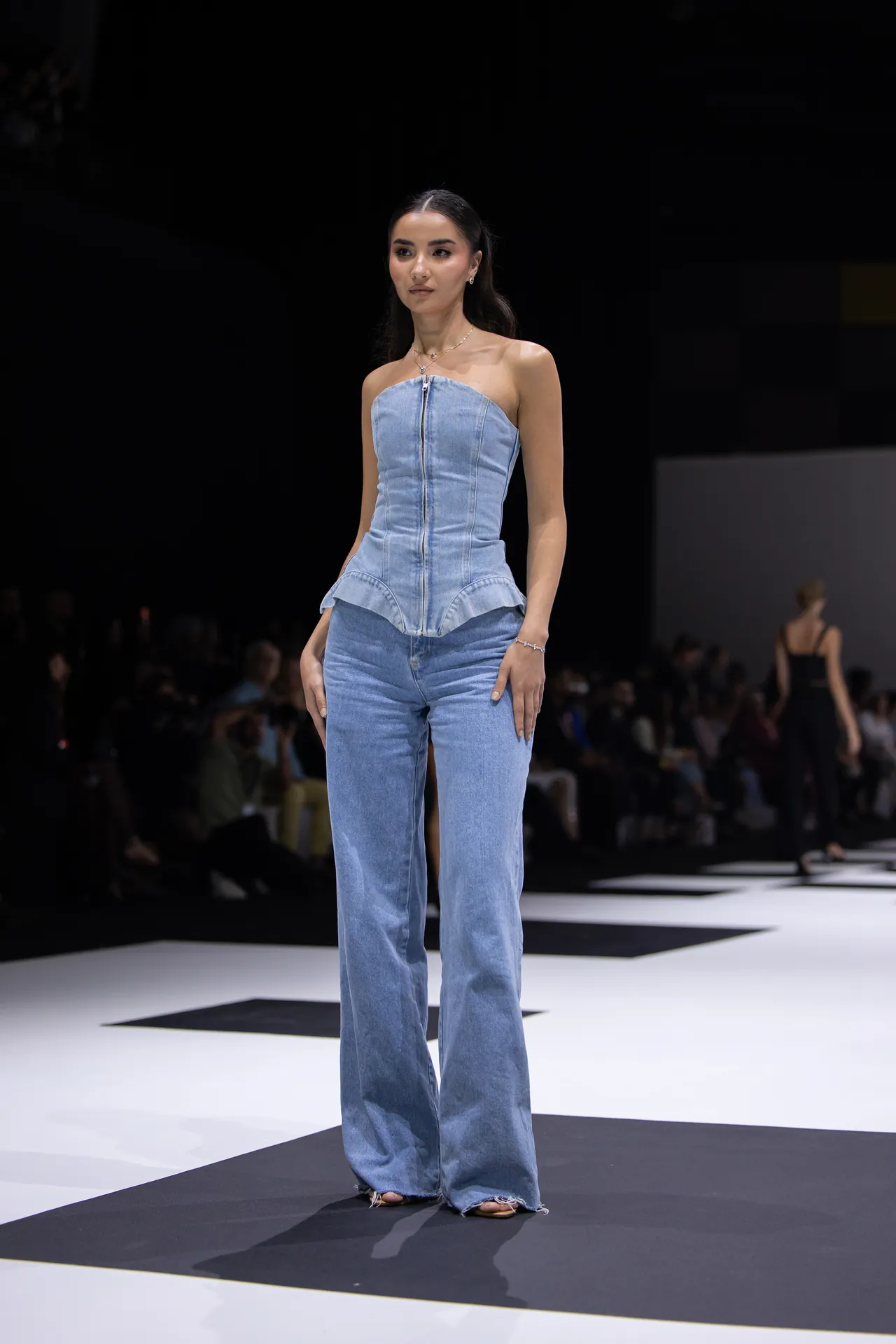 Female model walks the runway in a light blue denim strapless top with a front zipper and matching wide-leg jeans; audience in the dim background.