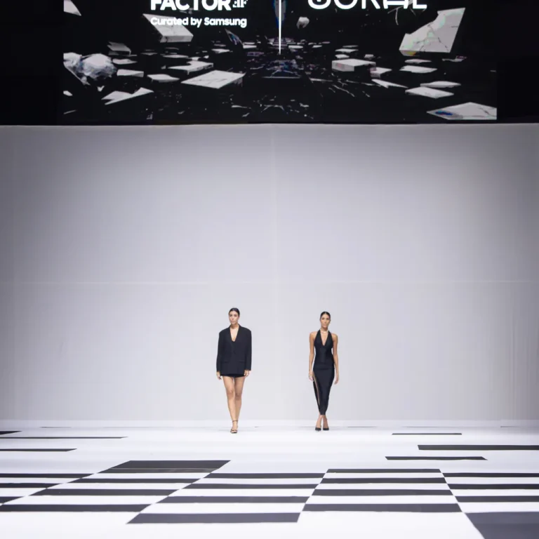 Two models walk a minimalist runway beneath a large screen showing Fashion Factor by Samsung and ZORAE logos behind them.