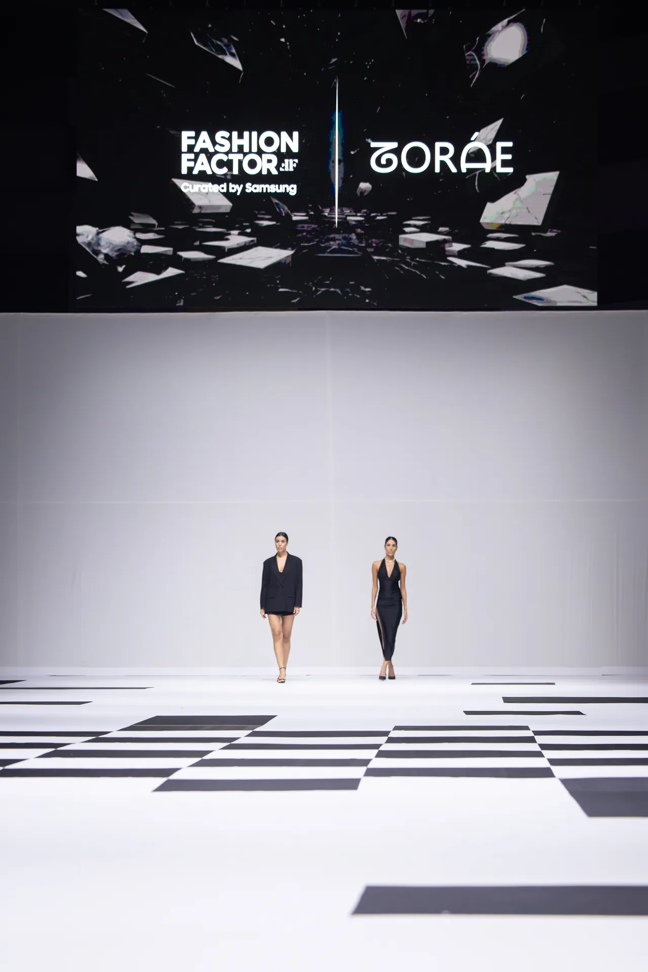 Two models walk a minimalist runway beneath a large screen showing Fashion Factor by Samsung and ZORAE logos behind them.