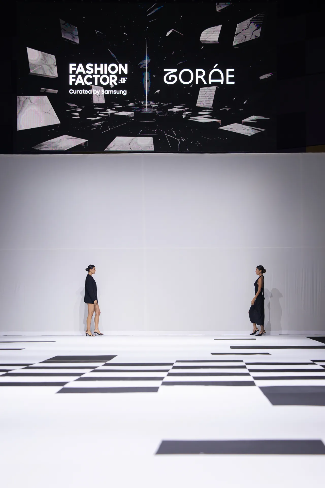 Two models walk on a white runway with a black geometric carpet while a large screen shows 'FASHION FACTOR' and designer name in the background.