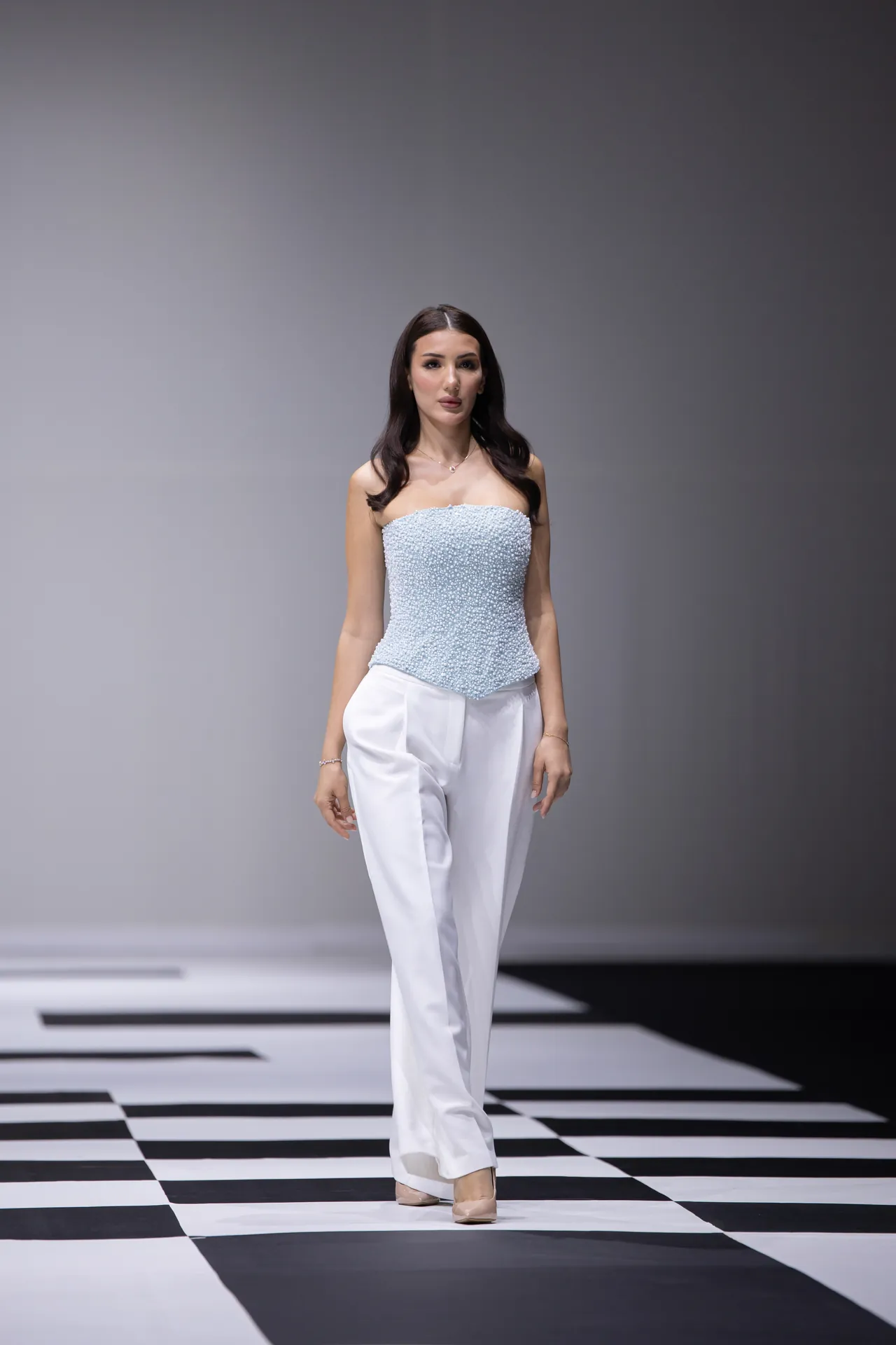 Female model walking a runway in a light blue beaded strapless top and white pants on a black-and-white checkerboard catwalk.