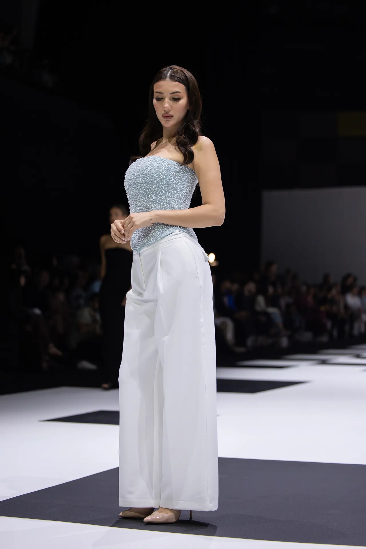 Model walking a runway in a light blue beaded strapless top and white wide-leg pants, beige heels, against a dark backdrop with an audience in the background.