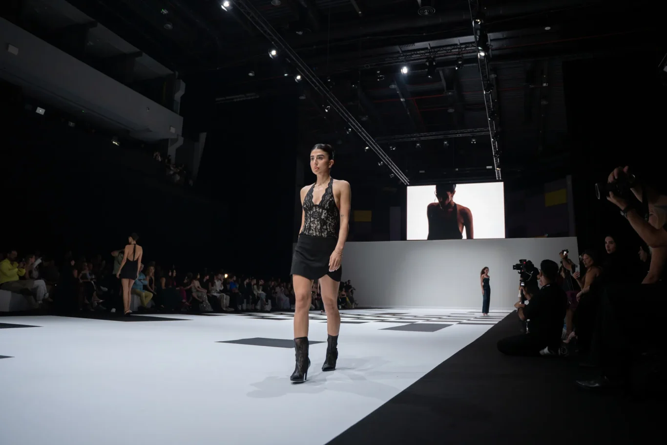 Model walks the fashion runway in a black lace top and mini skirt, knee-high boots, with audience and bright stage lights in the background.