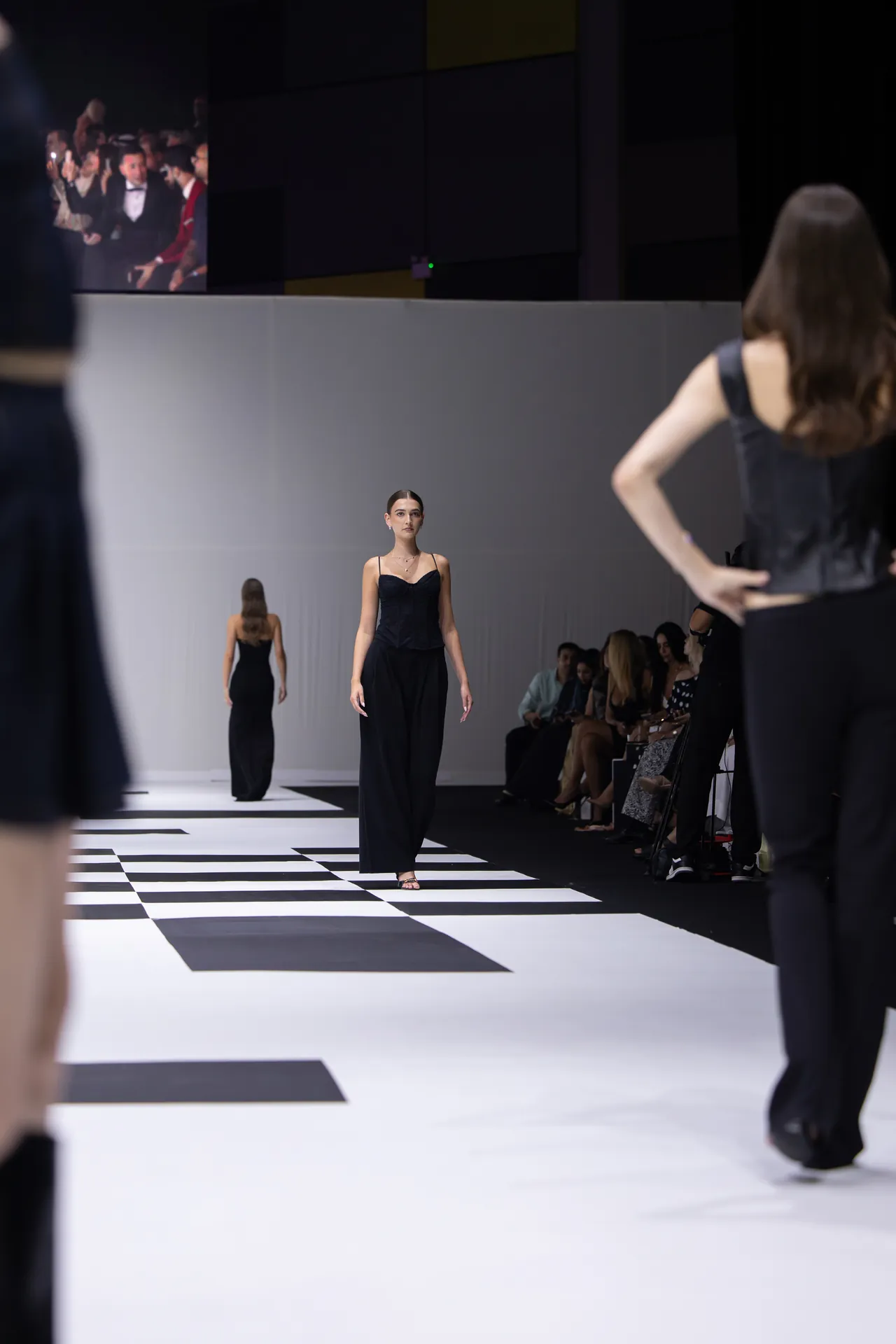 Model in black jumpsuit walks down a black-and-white checkered runway as seated audience watches on the right.