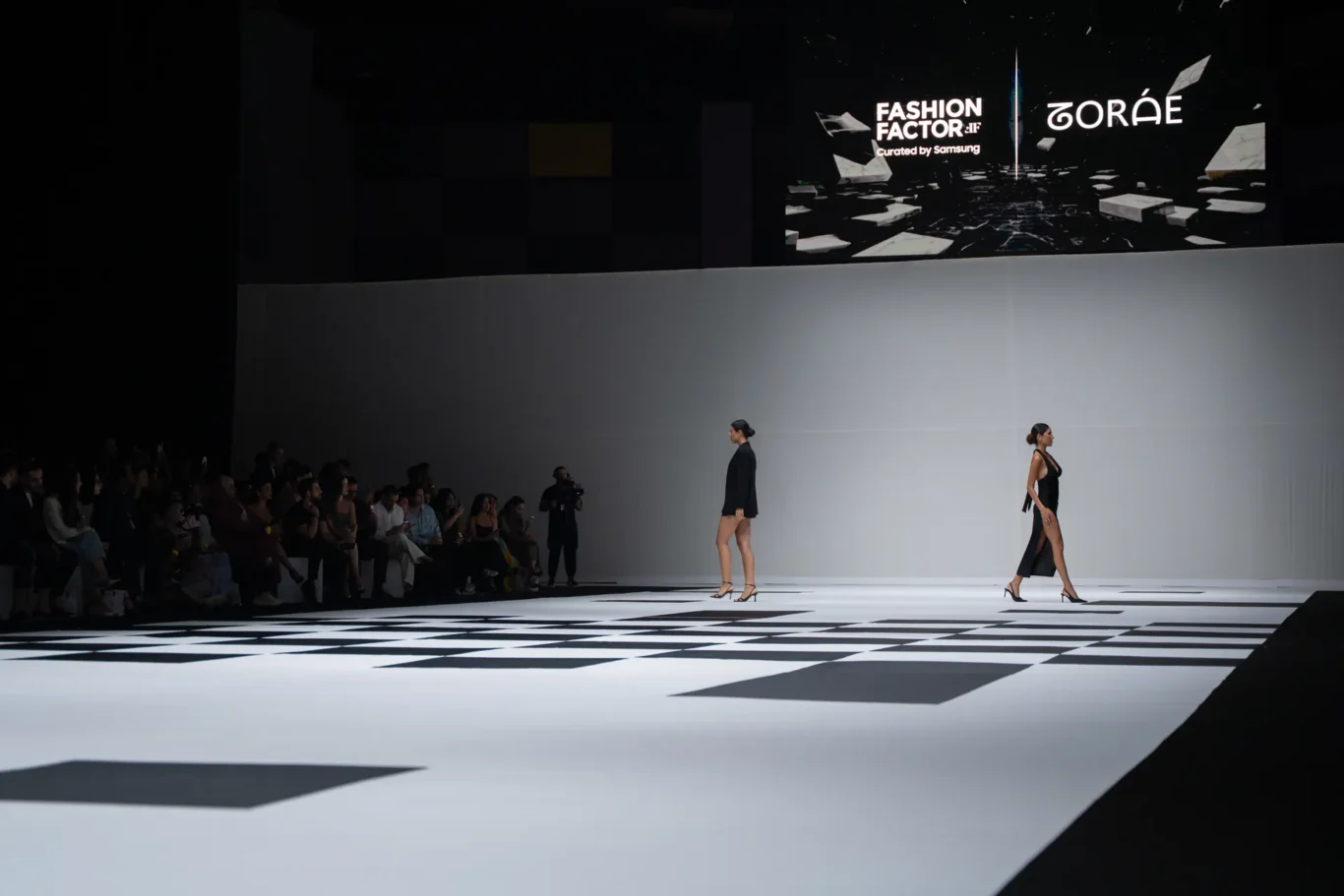 Two models walk a white runway with a bold black-and-white geometric floor as an audience watches from the left; large screens show logos in the background.
