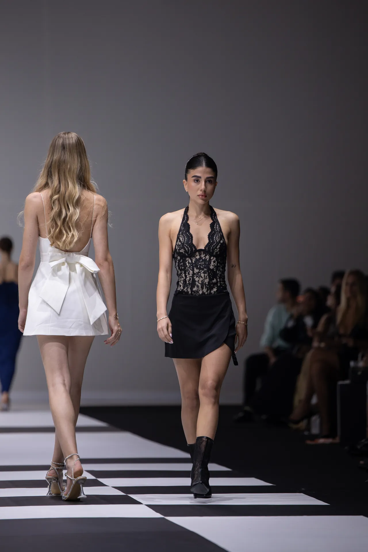 Two female models walk a fashion runway; the foreground model wears a black lace halter bodice and short skirt, the other wears a white backless dress seen from behind.