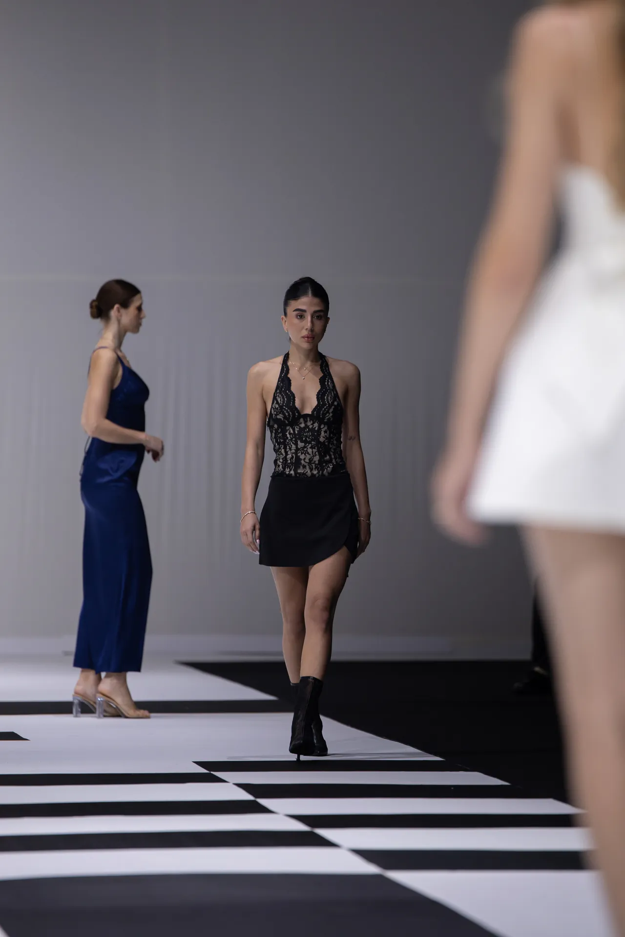 Model walks the runway in a black lace halter top and short black skirt, with other models in the background on a striped catwalk.