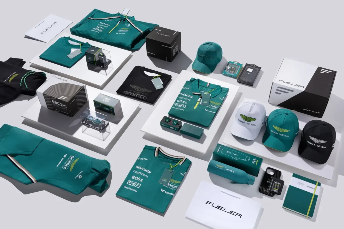 Collection of teal and black Formula One team merchandise displayed on white platforms, including hats, shirts, bags, and boxes.