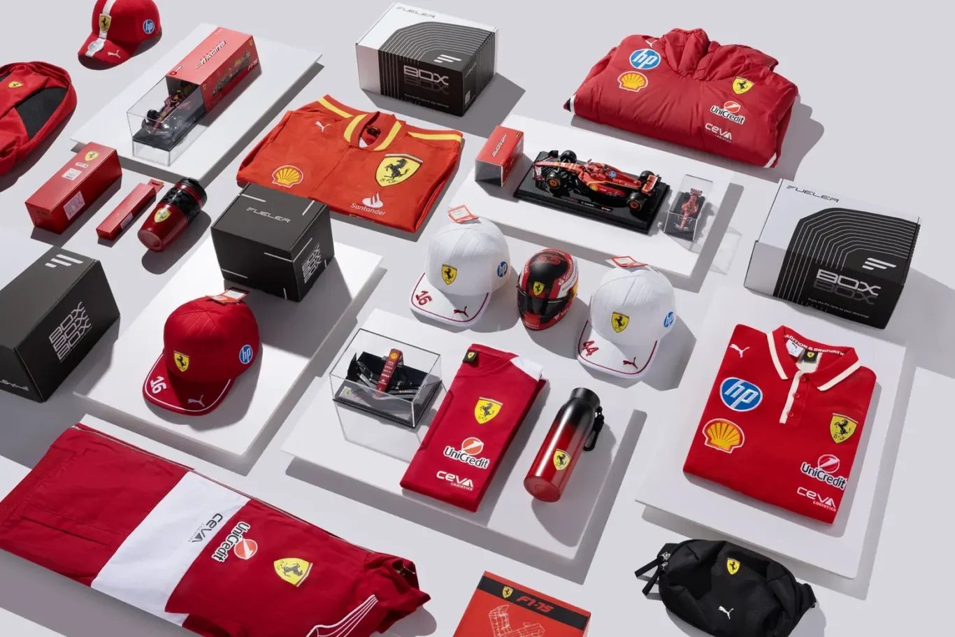 Ferrari-branded merchandise displayed on white platforms, including red shirts, caps, water bottles, and boxed gear with sponsor logos like Shell and HP.