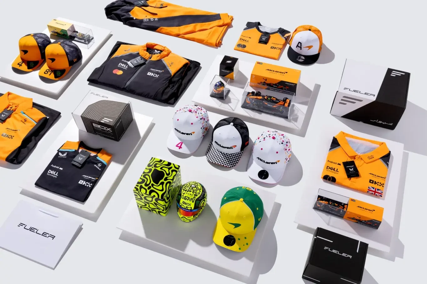 McLaren-branded merchandise laid out on white display platforms, including orange and black jackets, hats, caps, and boxed collectibles.