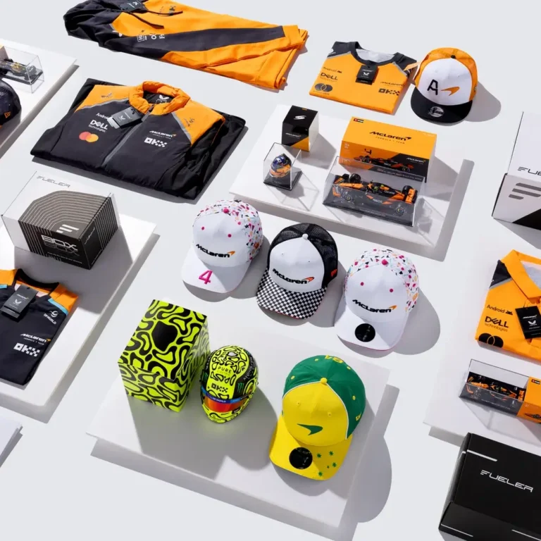 McLaren-branded merchandise laid out on white display platforms, including orange and black jackets, hats, caps, and boxed collectibles.