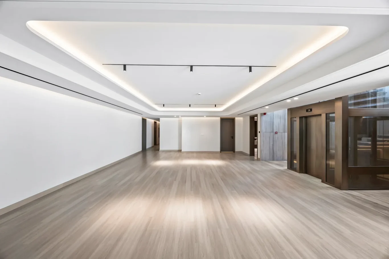 Spacious, modern lobby with light wood flooring, white walls, and recessed ceiling lighting; glass elevator banks on the right side.