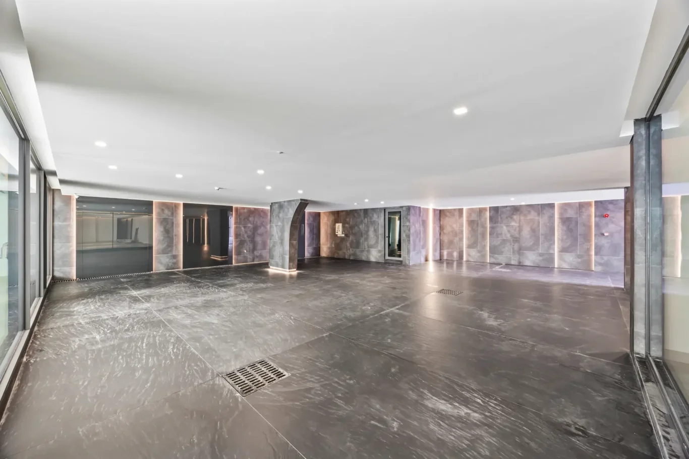 Empty modern lobby with dark marble floors, gray marble walls, and glass doors on the left beneath recessed lighting.
