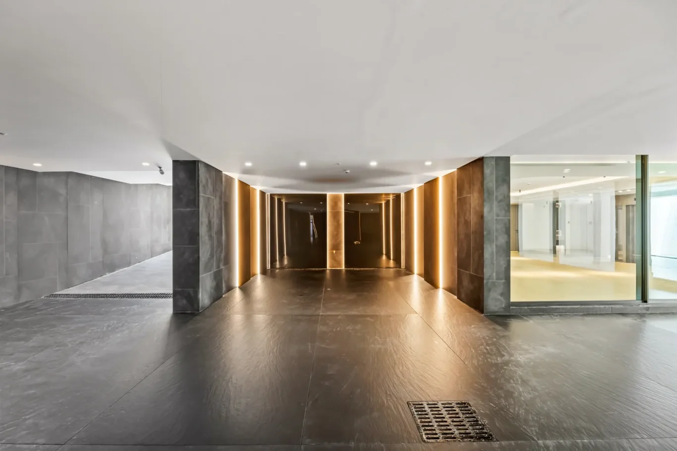 Modern indoor corridor: a dark marble tunnel with warm vertical light strips along the walls, leading to a brighter area ahead.