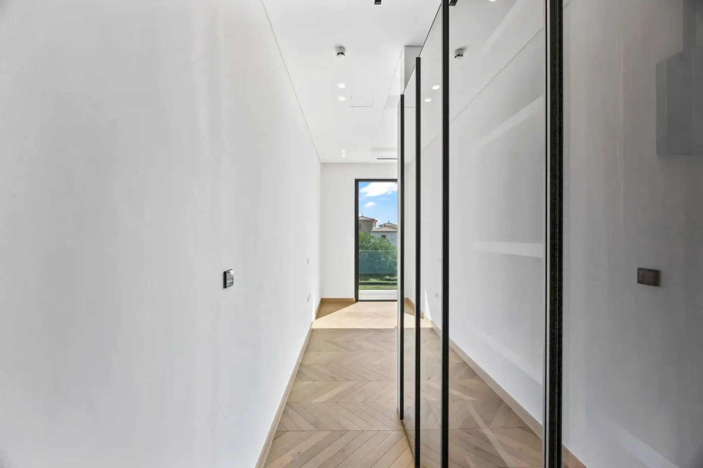 Narrow white-walled hallway with light wood herringbone floor and tall black-framed glass panels along the right, leading to a window showing blue sky and greenery.