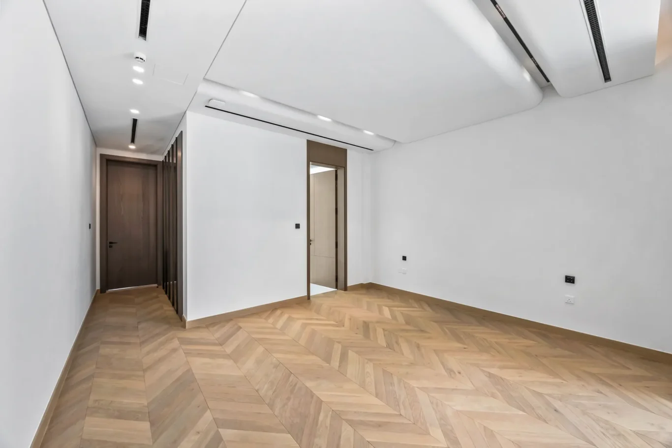 Bright hallway with white walls, dark wooden doors along the left, and a light herringbone wood floor.