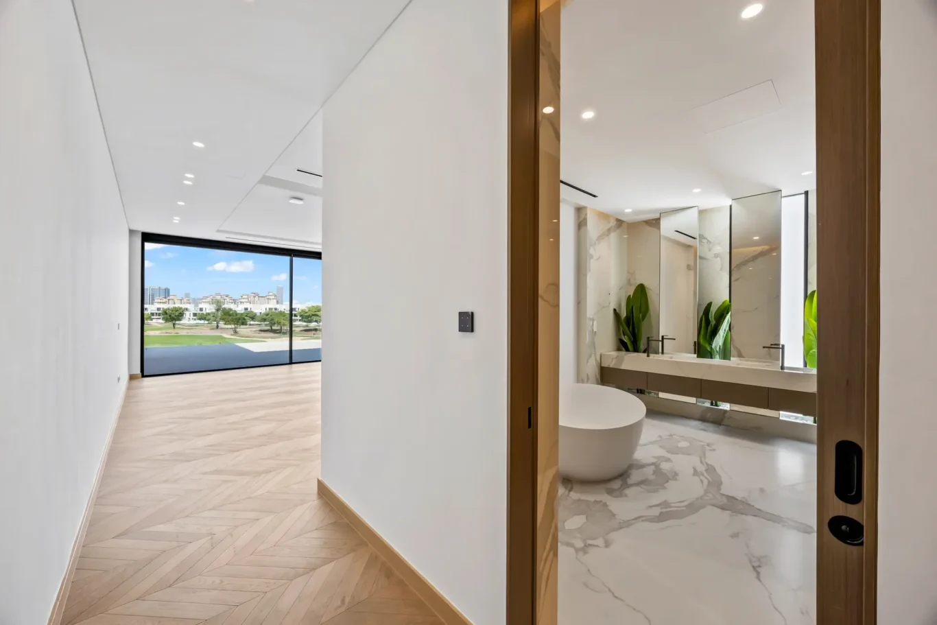 Bright open hallway with a herringbone wood floor leading to a marble bathroom, featuring a freestanding white tub, large mirrors, and greenery.