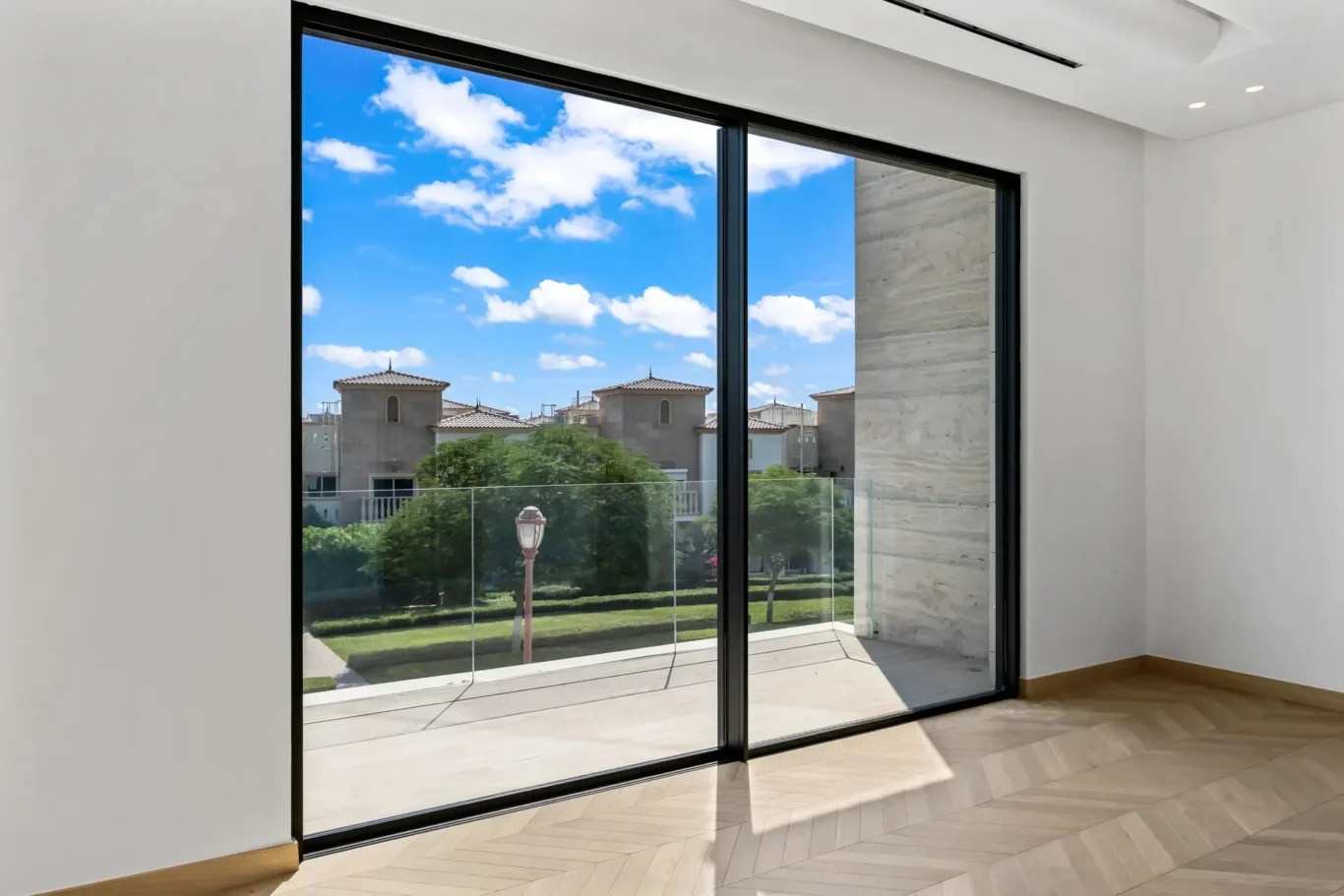 Interior room with a large glass sliding door opening to a balcony; view of suburban houses, trees, and a bright blue sky outside.