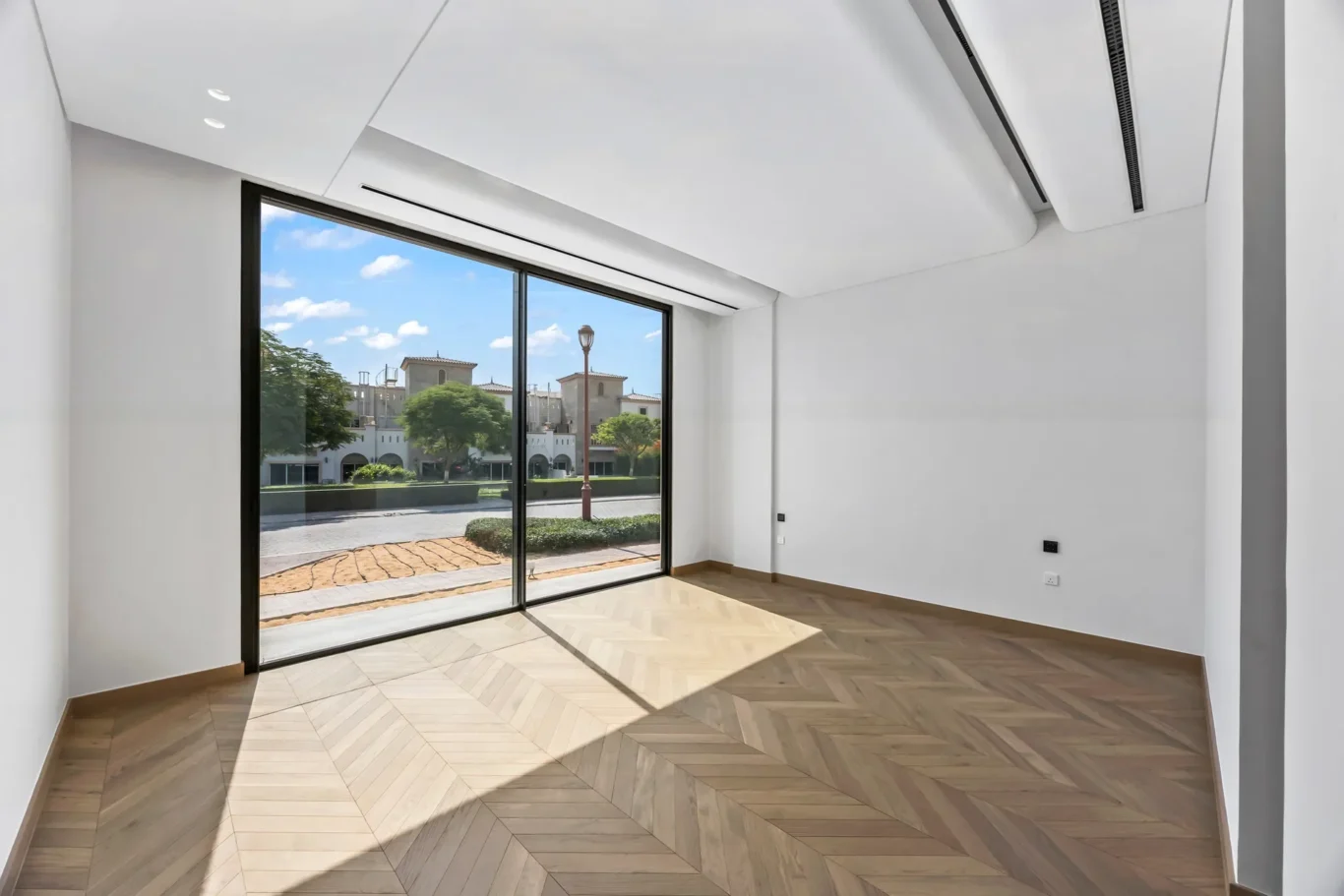 Bright empty room with a large floor-to-ceiling window showing the street and buildings outside, with sunlight on the floor.