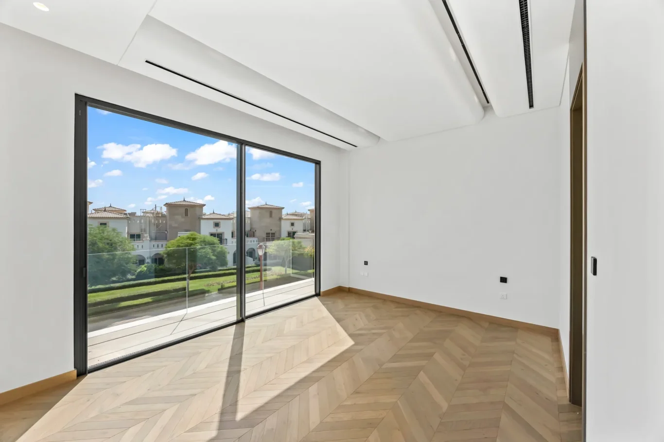 Bright empty living room with large sliding glass doors to a balcony and a sunny neighborhood view, herringbone wood floor.