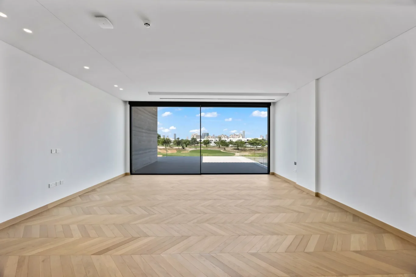 Empty bright room with herringbone wooden floor and large glass sliding doors opening to a park view and city skyline outside