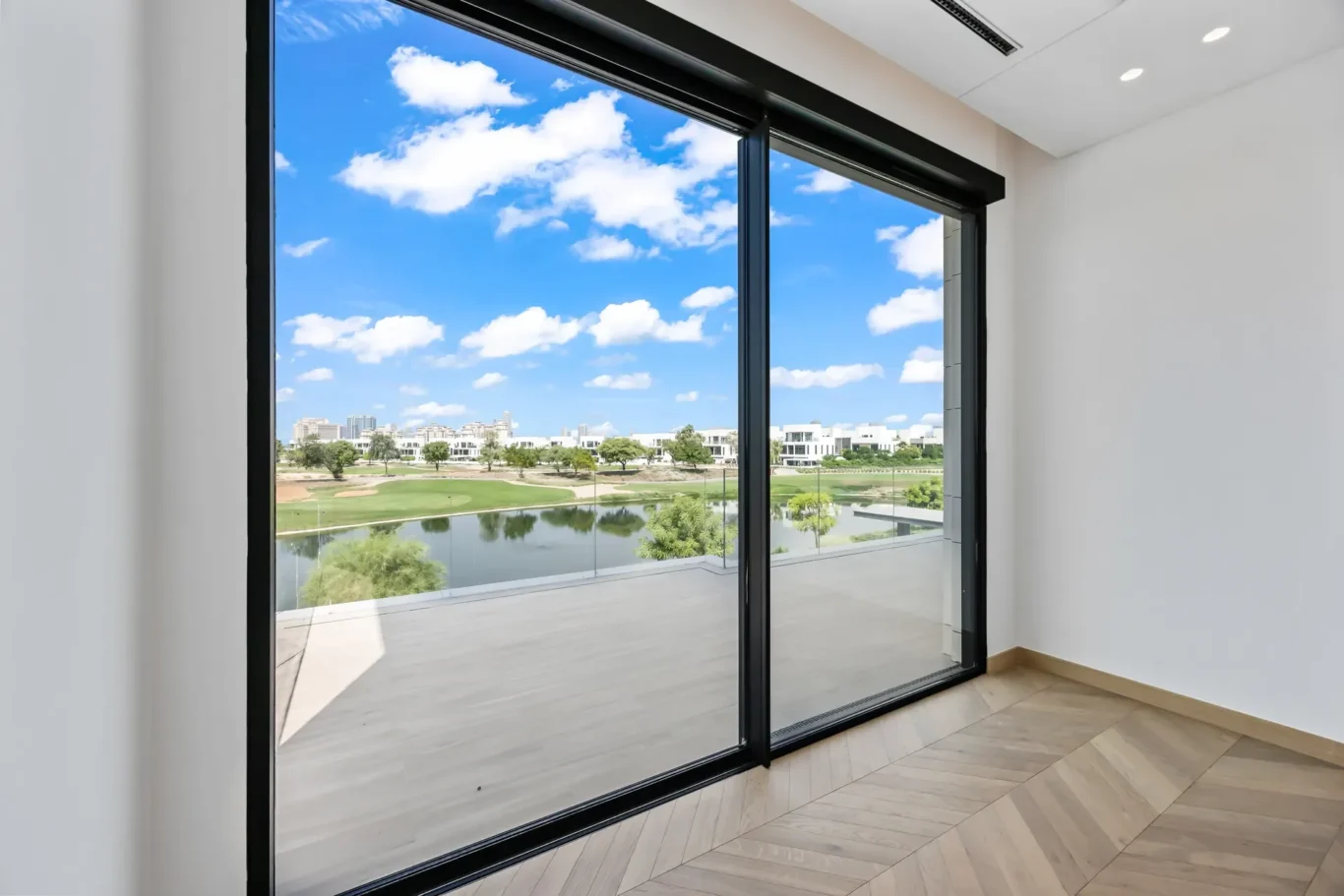 Interior with large glass sliding doors opening to a balcony; view of a park, pond, and distant buildings under a blue sky with clouds.