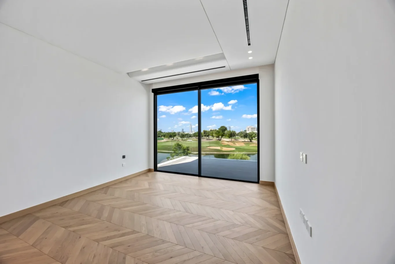 Empty, bright minimalist room with chevron wood flooring and a large glass sliding door opening to a sunny outdoor landscape.