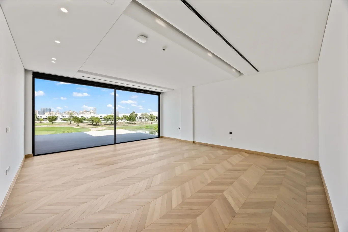 Empty living room with chevron wood floor and a large sliding glass door to a balcony overlooking the city park and buildings beyond.