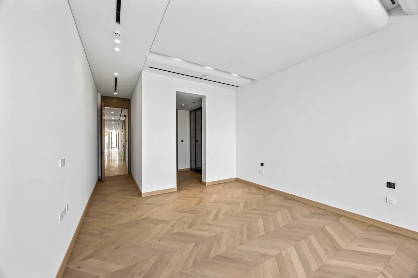 Empty modern hallway with white walls, a herringbone wood floor, and ceiling lights