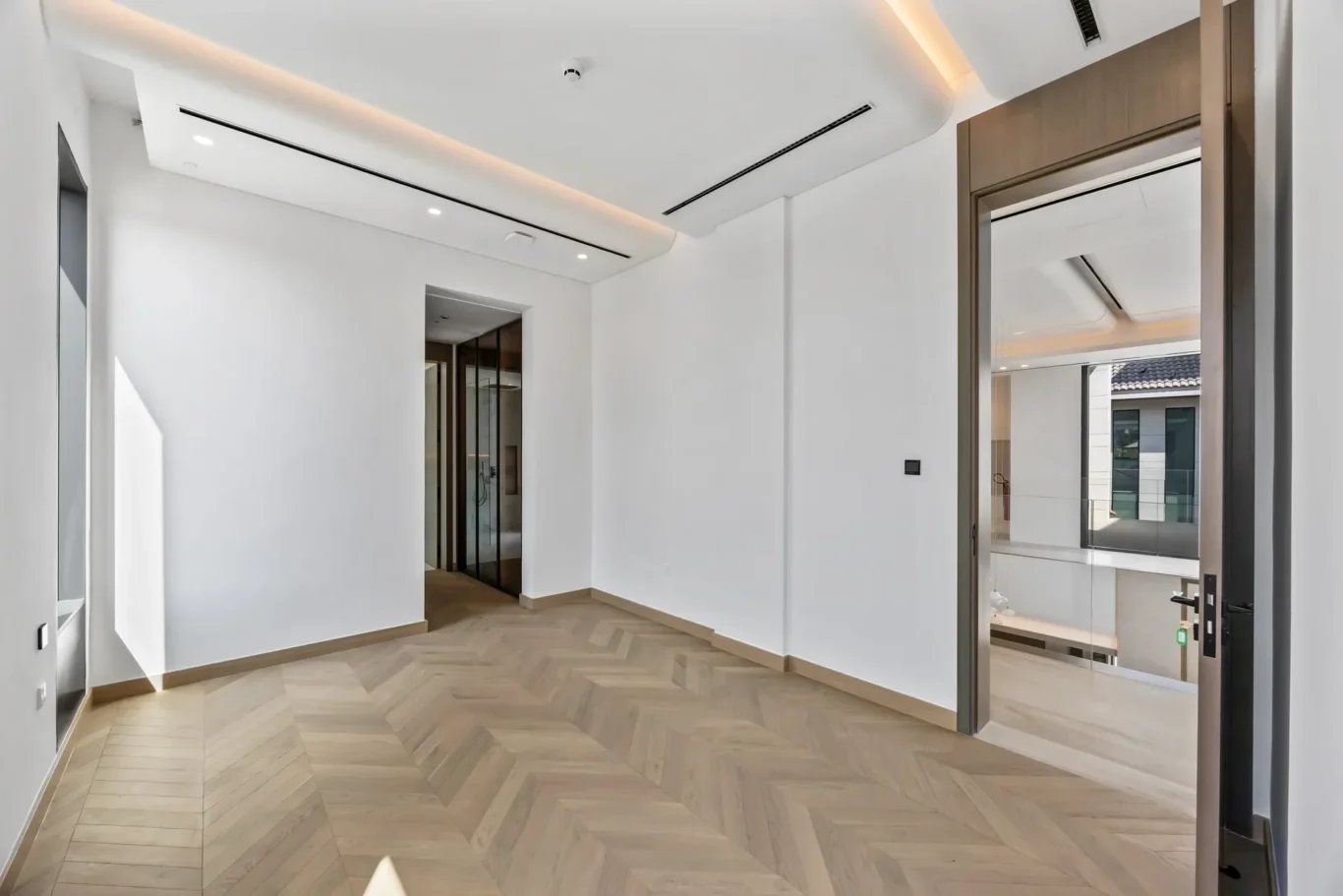 Bright modern hallway with white walls, herringbone wood floor, and glass doors to an exterior balcony.